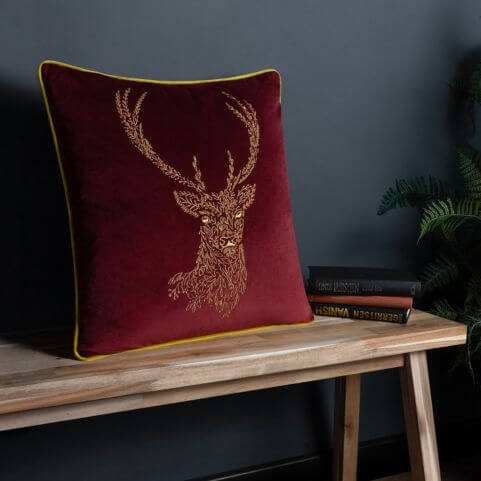 A burgundy and gold coloured velvet cushion with an embroidered woodland design of a stag, resting on a wooden bench in front of a blue background.
