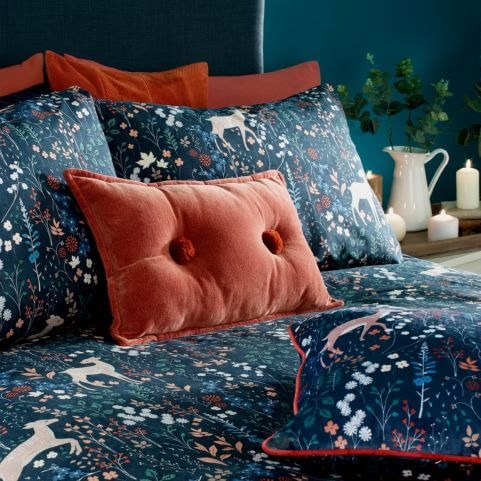 A midnight blue duvet cover set with a nature-inspired design of fallow deer and woodland florals, made on a bed with a complementary red scatter cushion.