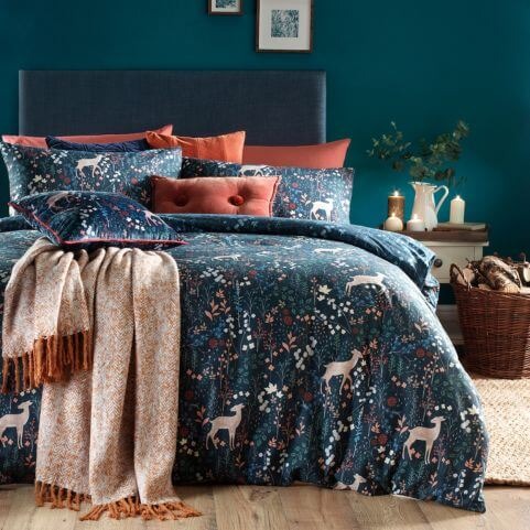 A midnight blue duvet cover set with a nature-inspired design of fallow deer and woodland florals, made on a bed with a complementary scatter cushions and a gold woven throw, presented in a bedroom with teal walls.