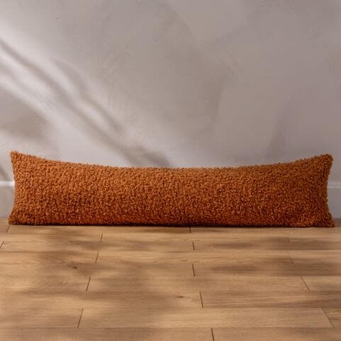 A ginger coloured draught excluder cushion with a chunky bouclé design, laid on a wooden floor in front of a white background.