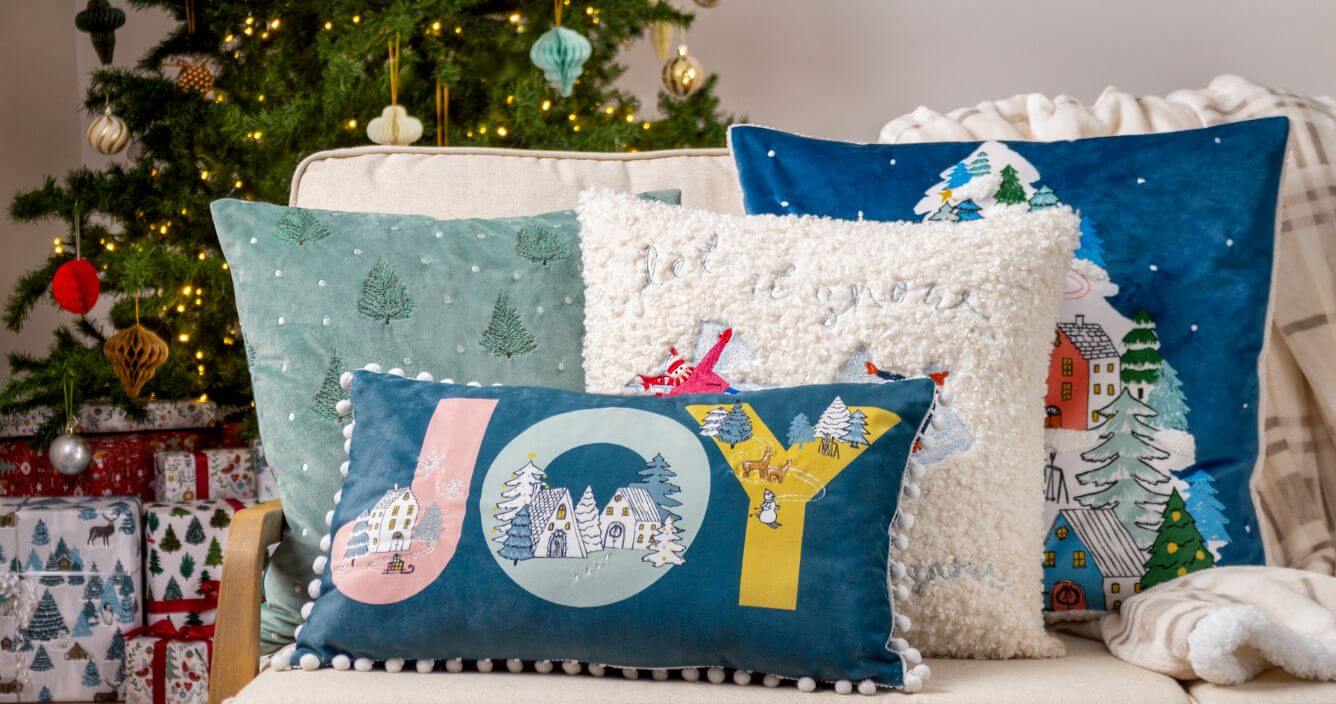 A selection of four Christmas cushions in neutral, blue and green shades, arranged on a cream sofa along with a cream fleece throw in front of a decorated Christmas tree.