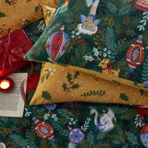 A green Christmas duvet cover set with a traditional design of baubles and other trinkets, arranged on a bed with a book, candle and christmas gifts.