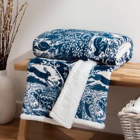 A midnight blue fleece throw with a printed winter woodland design, folded on a wooden bench in front of a white background.