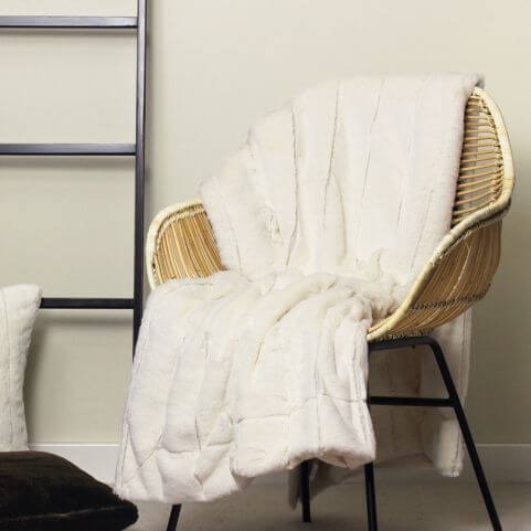 A cream-coloured faux fur throw blanket, draped over a woven chair next to coordinating furnishings in a neutral room.