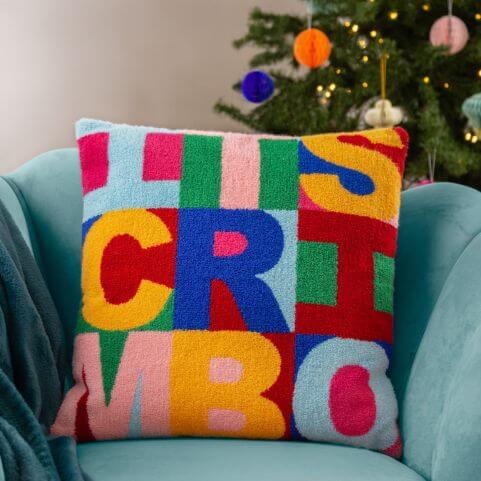 A knitted technicolour cushion in a woven loop cotton design, complete with a slogan reading 'it's Crimbo'.