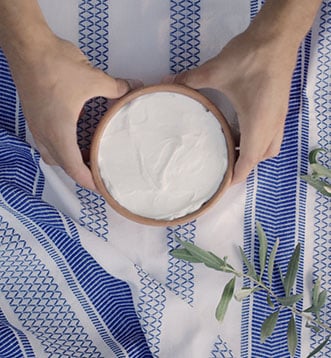 Zwei Hände halten Keramiktopf mit frischem griechischen Joghurt über einer blau-weißen Tischdecke im griechischen Folklore-Muster