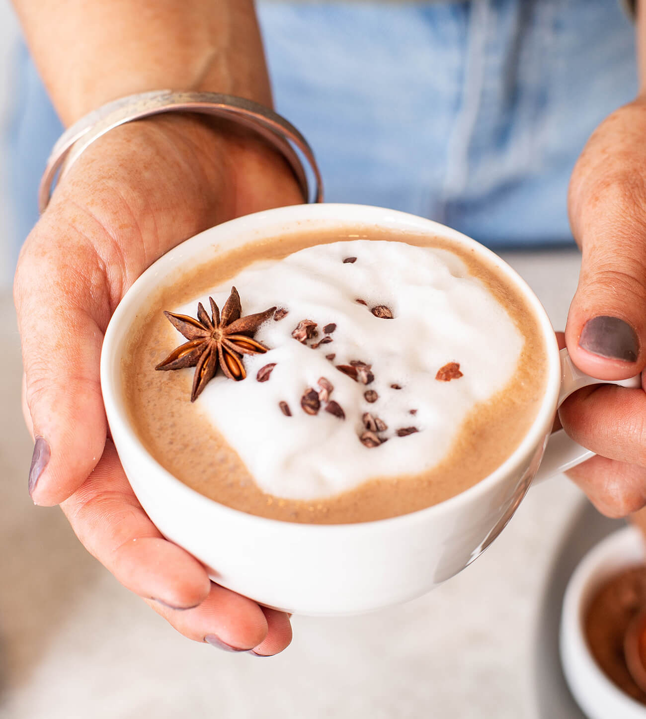 Holding a cup of Winter Chai Hot Chocolate with a topped with milk foam, chocolate shavings and an anise seed pod