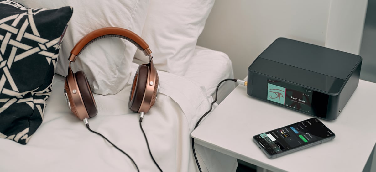 Headphones on a bed next to a night stand with a smart phone and streamer