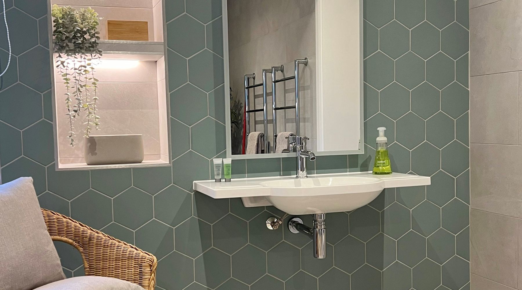 Attractive wall mounted basin and illuminated mirror with storage niche and wicker chair against hexagon tiles in Timeless Jade