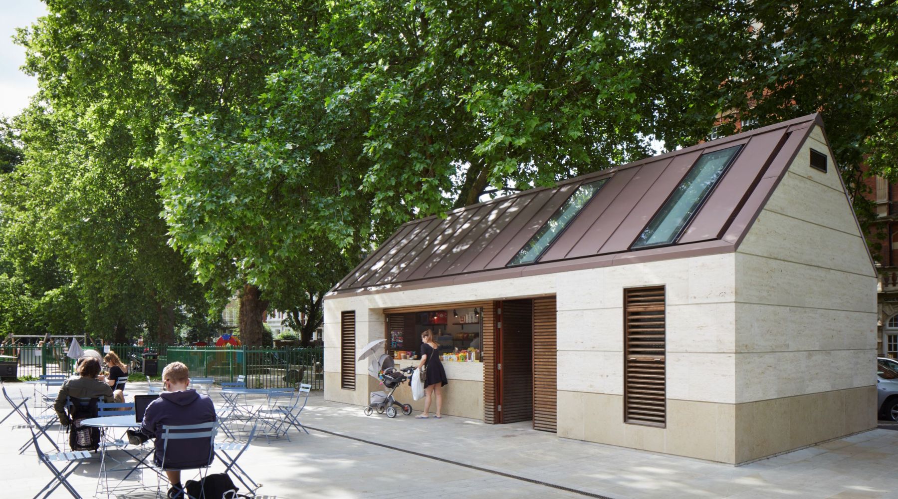 External shot of Brook Green Pavilion with leafy green trees behind and people sitting on outdoor furniture in front 