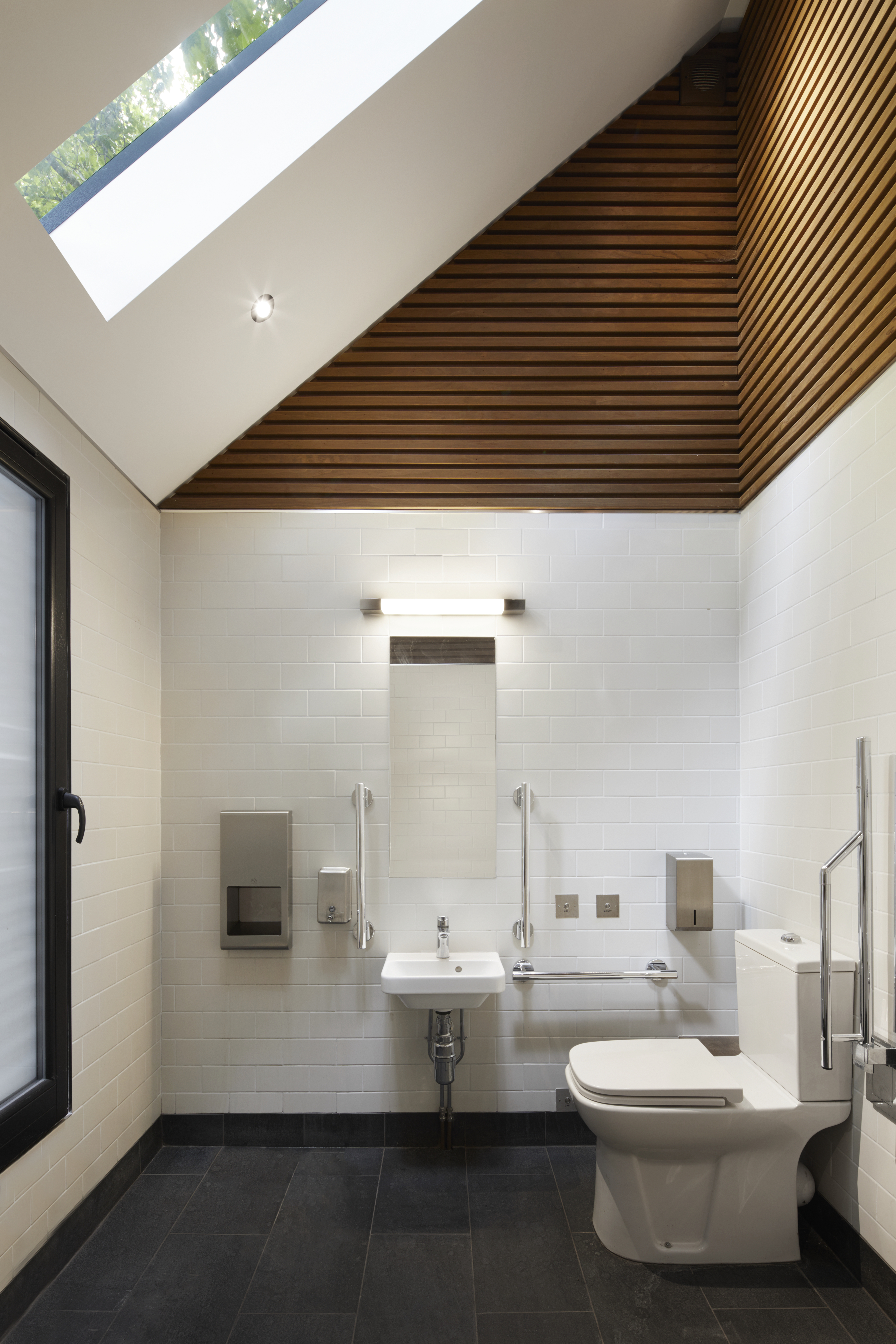 Brook Green Pavilion’s design-led Doc M compliant accessible toilet including wood panelled vaulted ceiling, chrome grab rails, raised height toilet with easy-to-use flush, and call alarm button. 