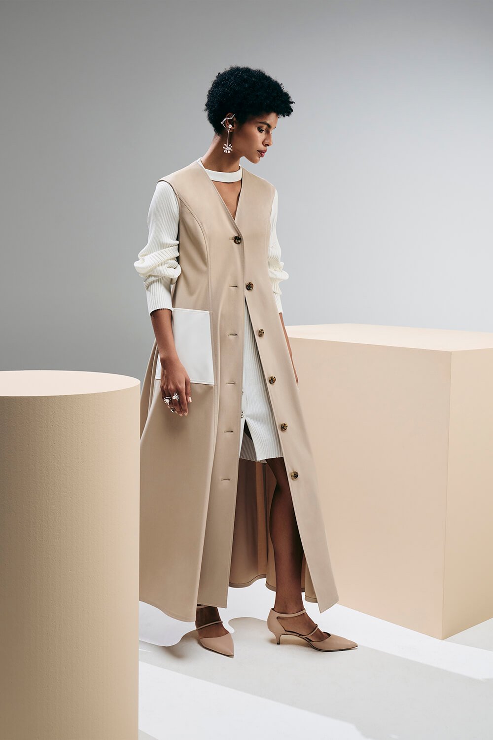 Model wears a sand trench coat with brown buttons over a white cardigan and white skirt