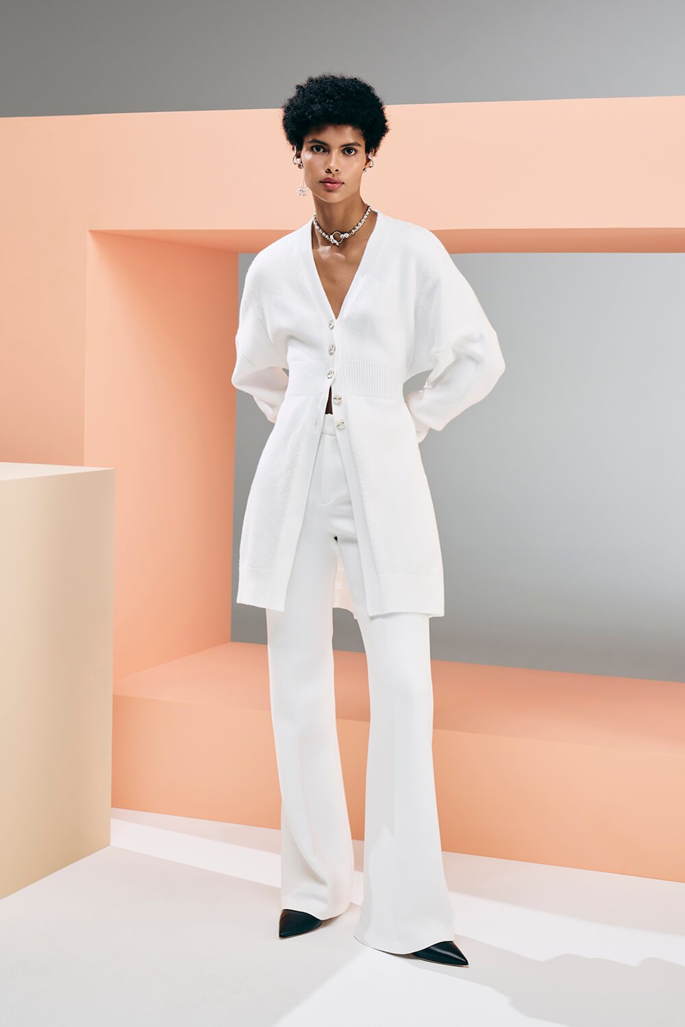 Model wears white flair trousers, with a white cardigan