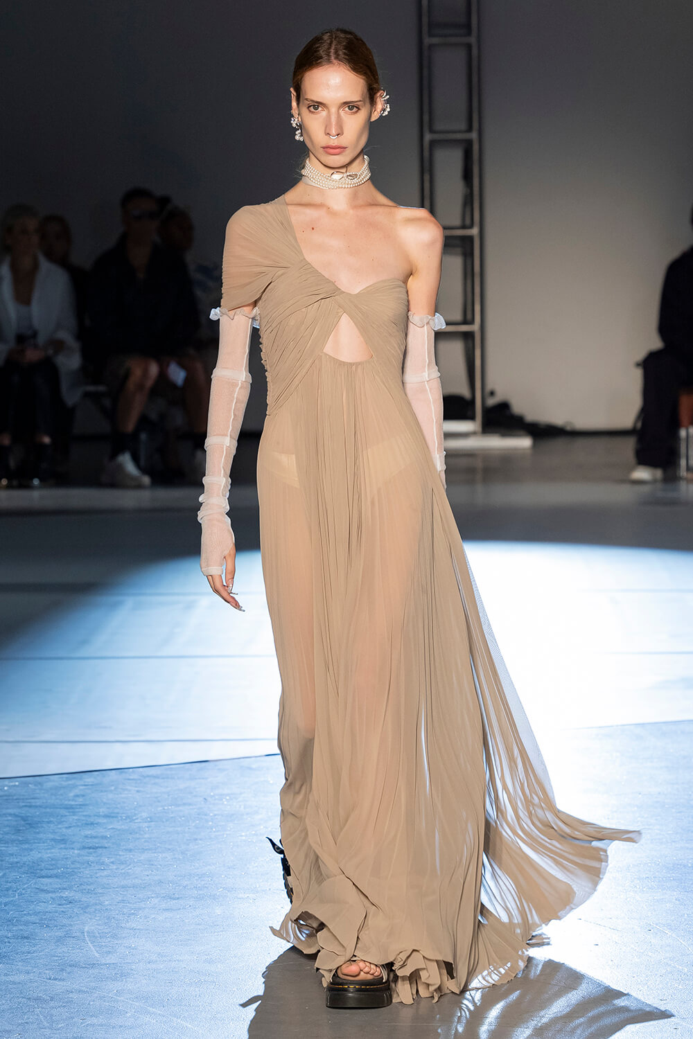 Model wears a sand colored long dress, see through with white gloves