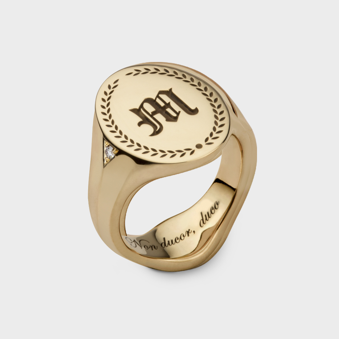 A yellow gold oval ring engraved with the letter M at the top and a pattern around it, with engraving on the inside bottom of the ring and a diamond on the side of the signet.