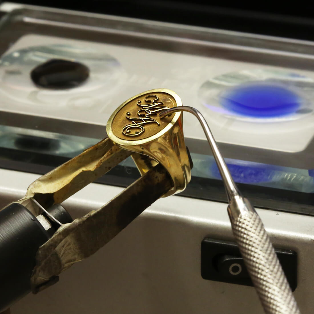Yellow gold oval signet ring with interlocking initials MAO in front of the enamelling section in the workshop ready to be enamelled with blue paint 