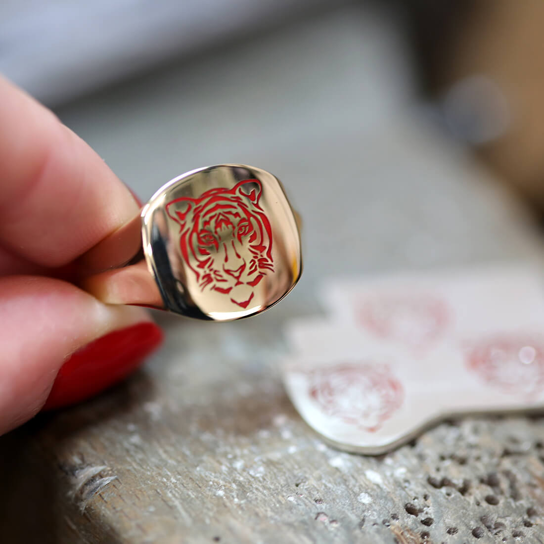 A jeweller with red nails holding a yellow gold signet ring with a red tiger enamelled on it.