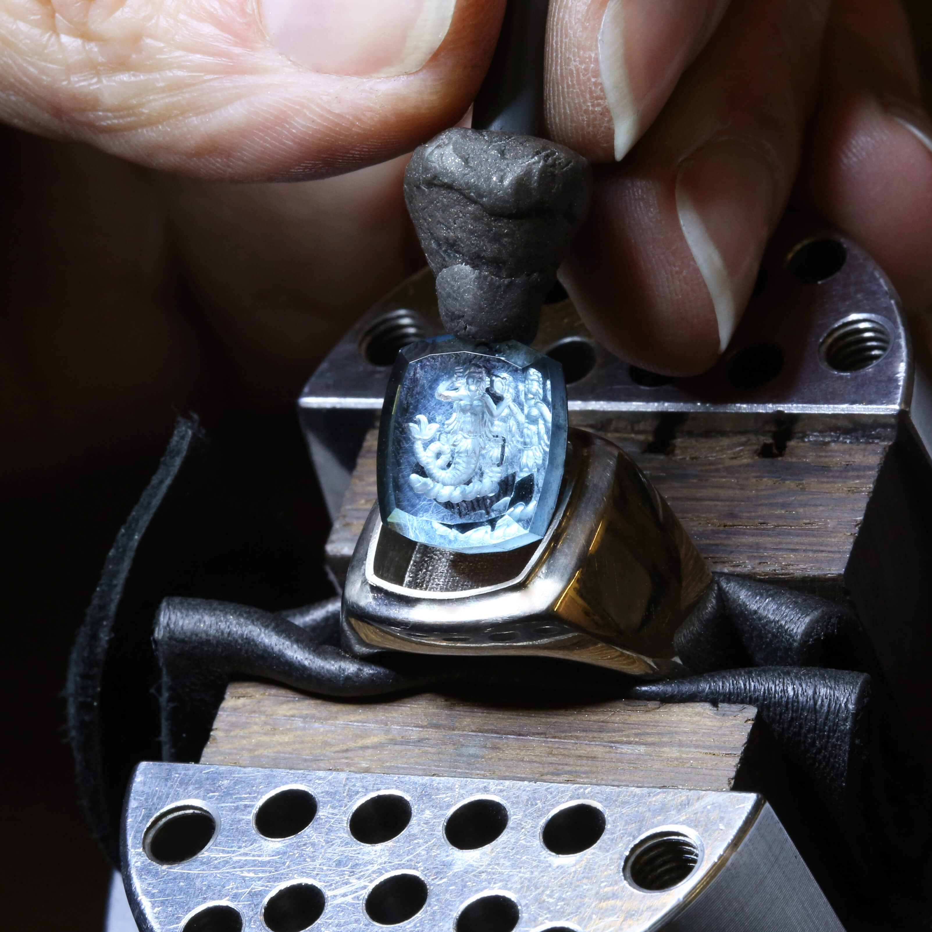A jeweller's hand in a workshop is setting a intaglio blue stone in a silver signet ring