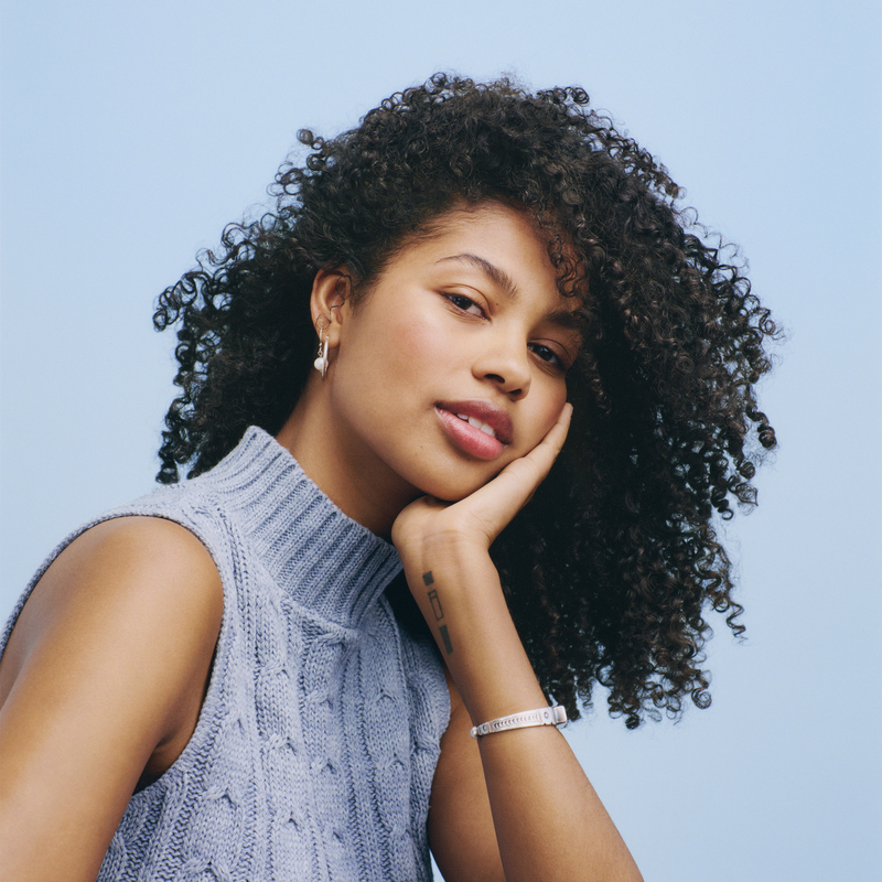 A person with curly hair, wearing a sleeveless knitted turtleneck, rests their head on their hand while gazing at the camera against a light blue background.