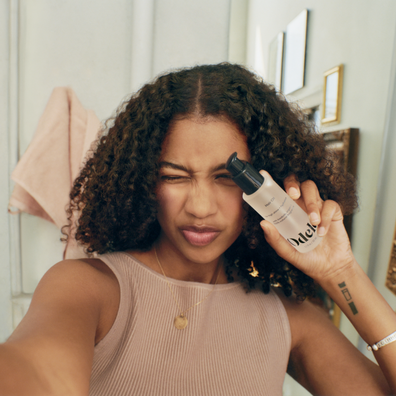 A person with curly hair holds up a bottle of Odele Hair Oil.