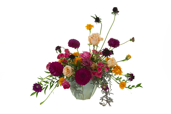 A vibrant and juicy-colored boho floral design mixes Italian ruscus, variegated pittosporum, acacia foliage, dahlias, marigolds, Blueberry roses, Shimmer roses, spray roses, ranunculus, and scabiosa in a pottery vessel.