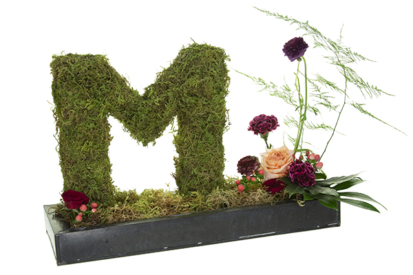 A lovely moss-covered monogram set into a base with beautiful blooms and foliage adds a unique touch to a guestbook or check-in table at a special event.