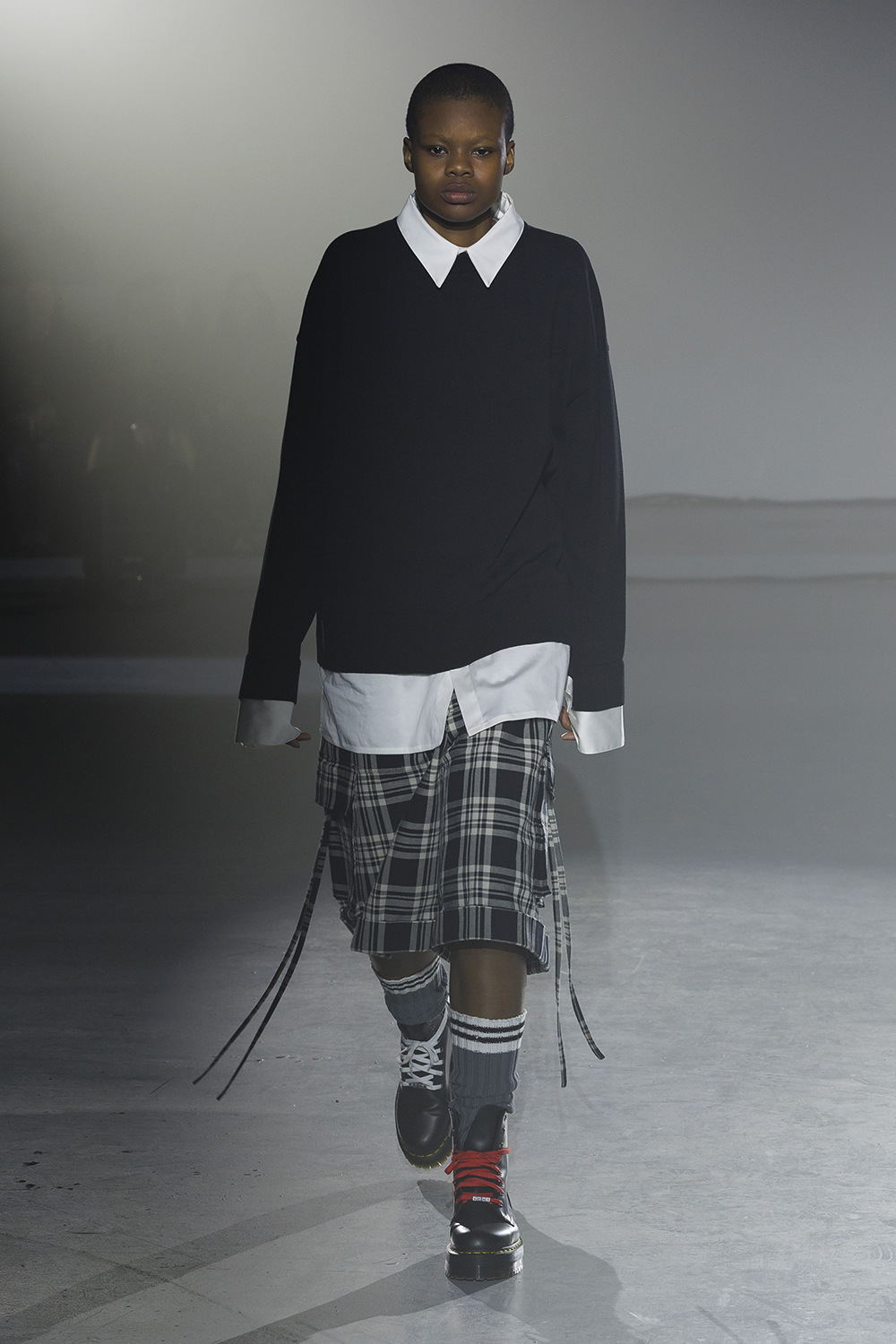 Model wears plaid knee length shorts and a navy sweater layered over a white button down