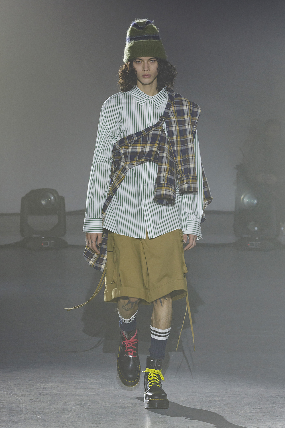 Model wears mustard knee length shorts with a green pinstripe button down and plaid jacket wrapped across body