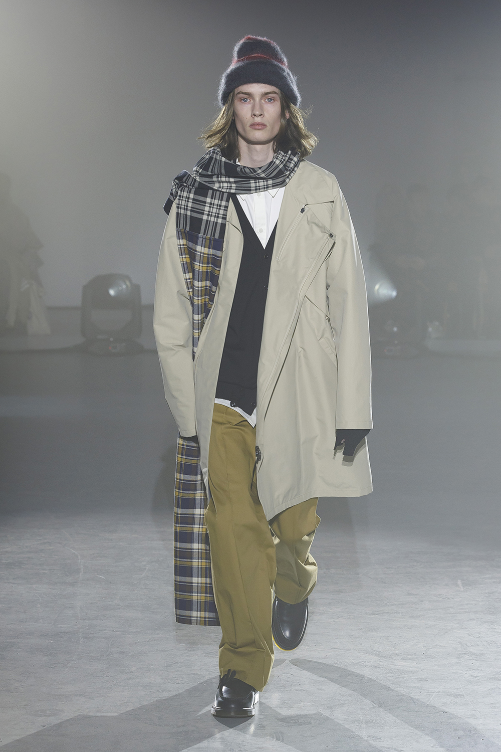 Model wears mustard pants, black v neck sweater over a white button down and layer with a cream trench coat and plaid scarf