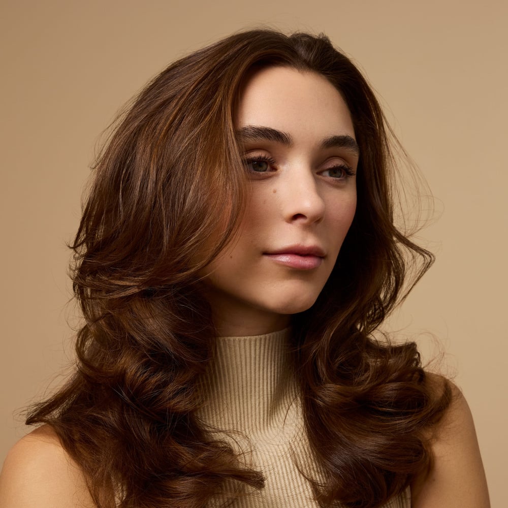 A woman with thick, healthy-looking wavy brown hair looks ahead. 