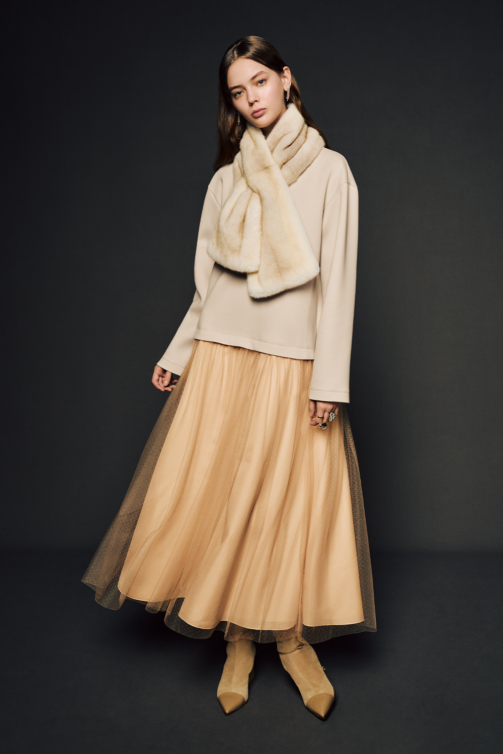 The model is wearing a beige long-sleeved top paired with a voluminous beige tulle skirt and a plush, oversized faux-fur scarf, completing the look with pointed beige ankle boots.