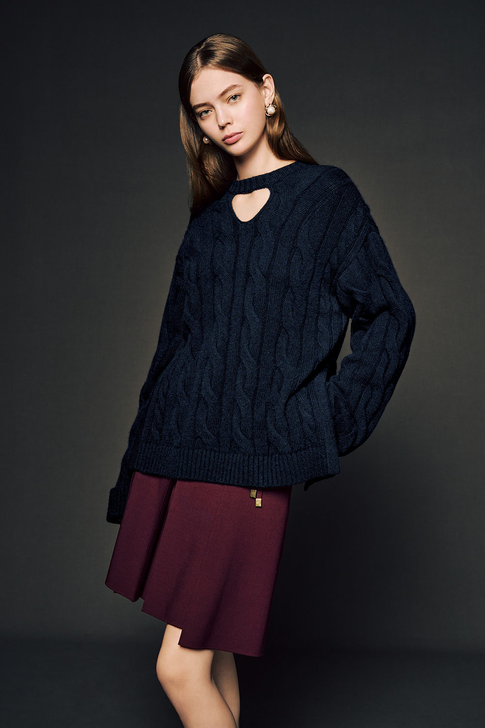 The model is wearing a navy blue cable-knit sweater with a small keyhole detail at the neckline, paired with a knee-length burgundy skirt.