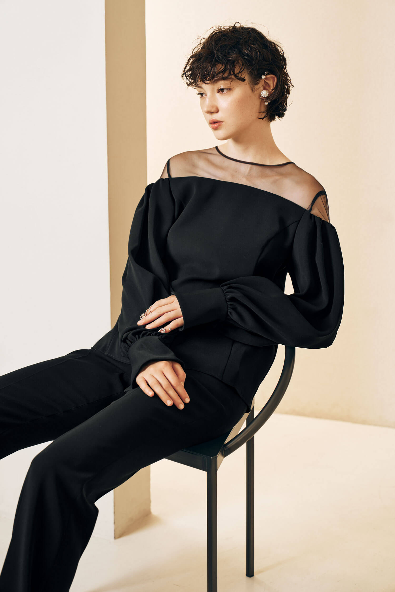 The model wears a sleek black ensemble featuring a long-sleeve top with sheer paneling at the shoulders and wide-leg pants. 