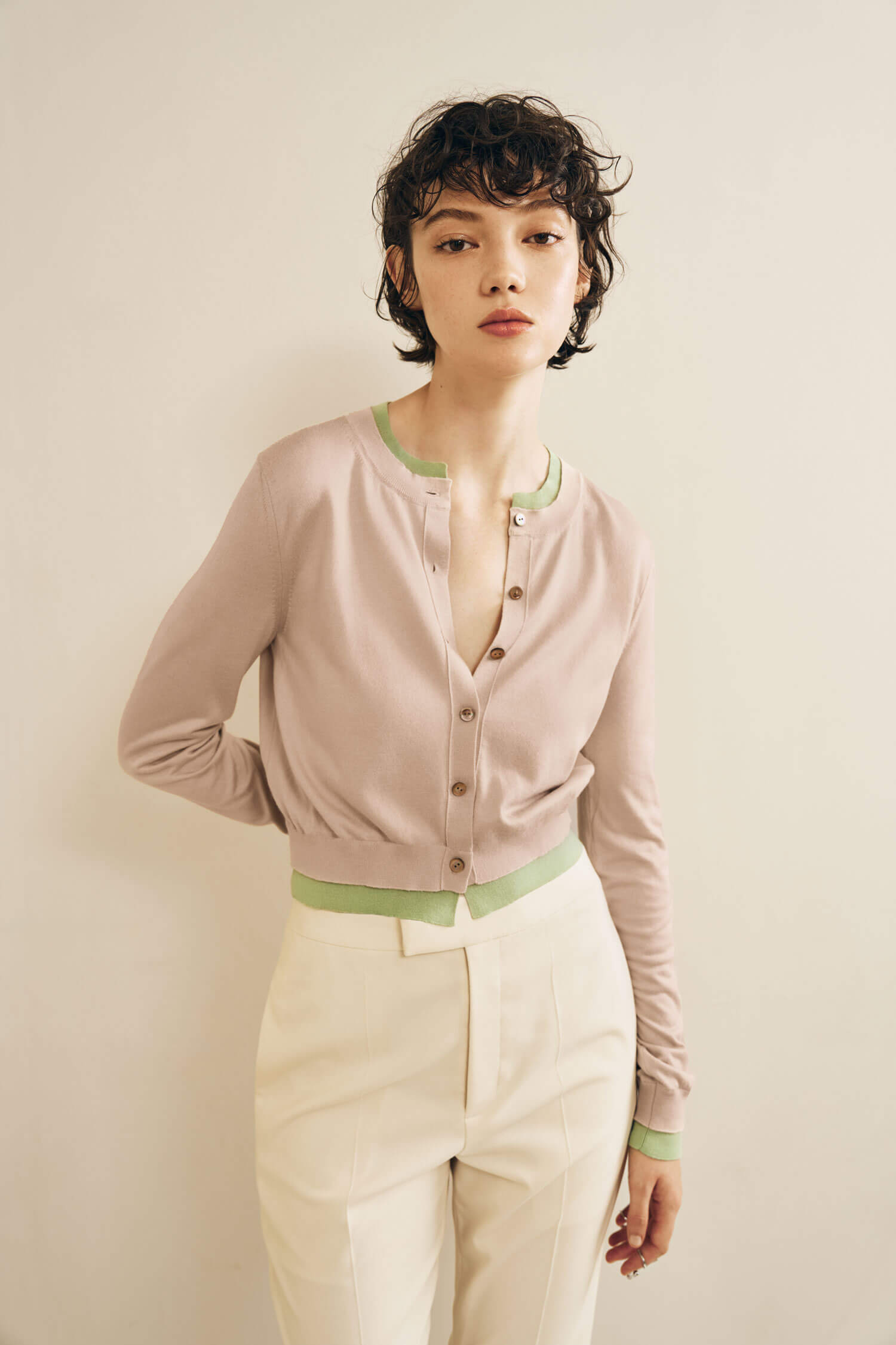 The model is wearing a light beige cardigan with green trim details, tucked into high-waisted cream trousers.
