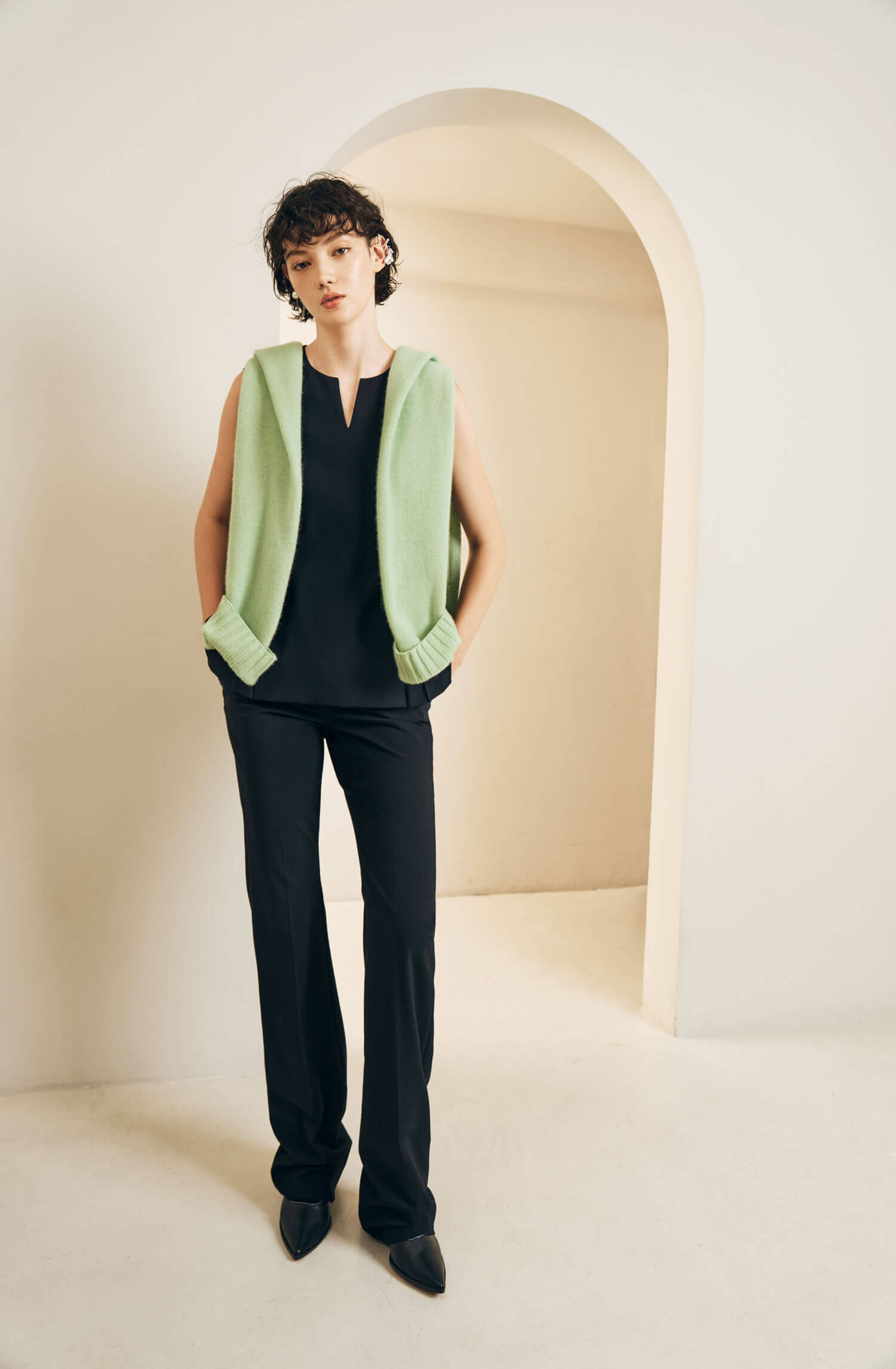 The model wears a black blouse and black trousers and a green sweater over the shoulders 