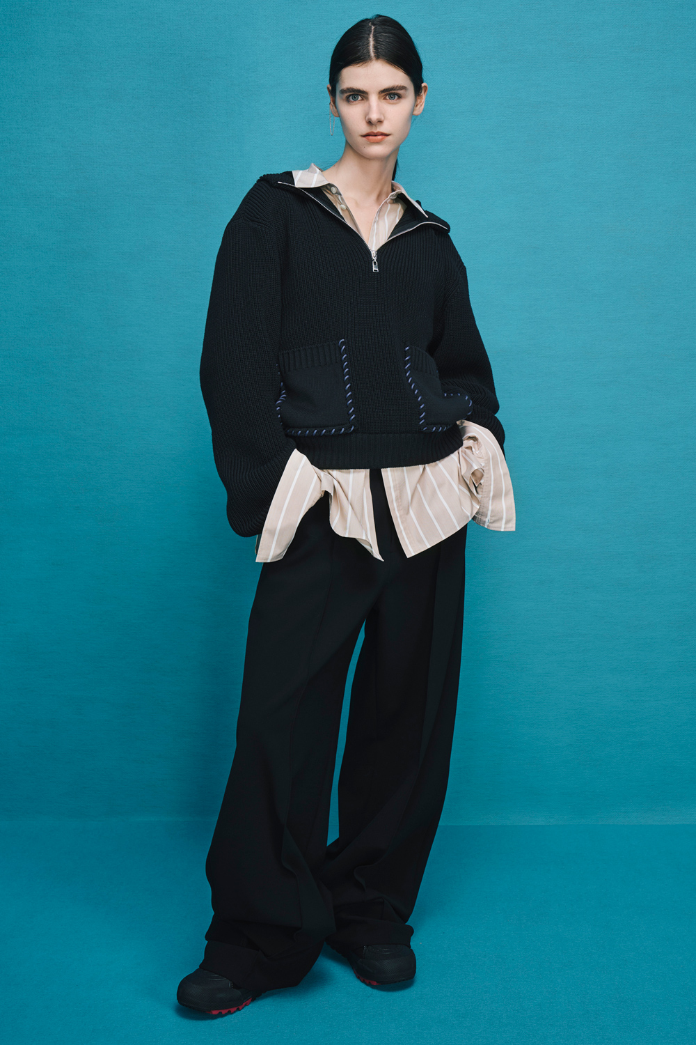 Model is wearing black pants with a black hoodie over a beige pinstripe shirt.