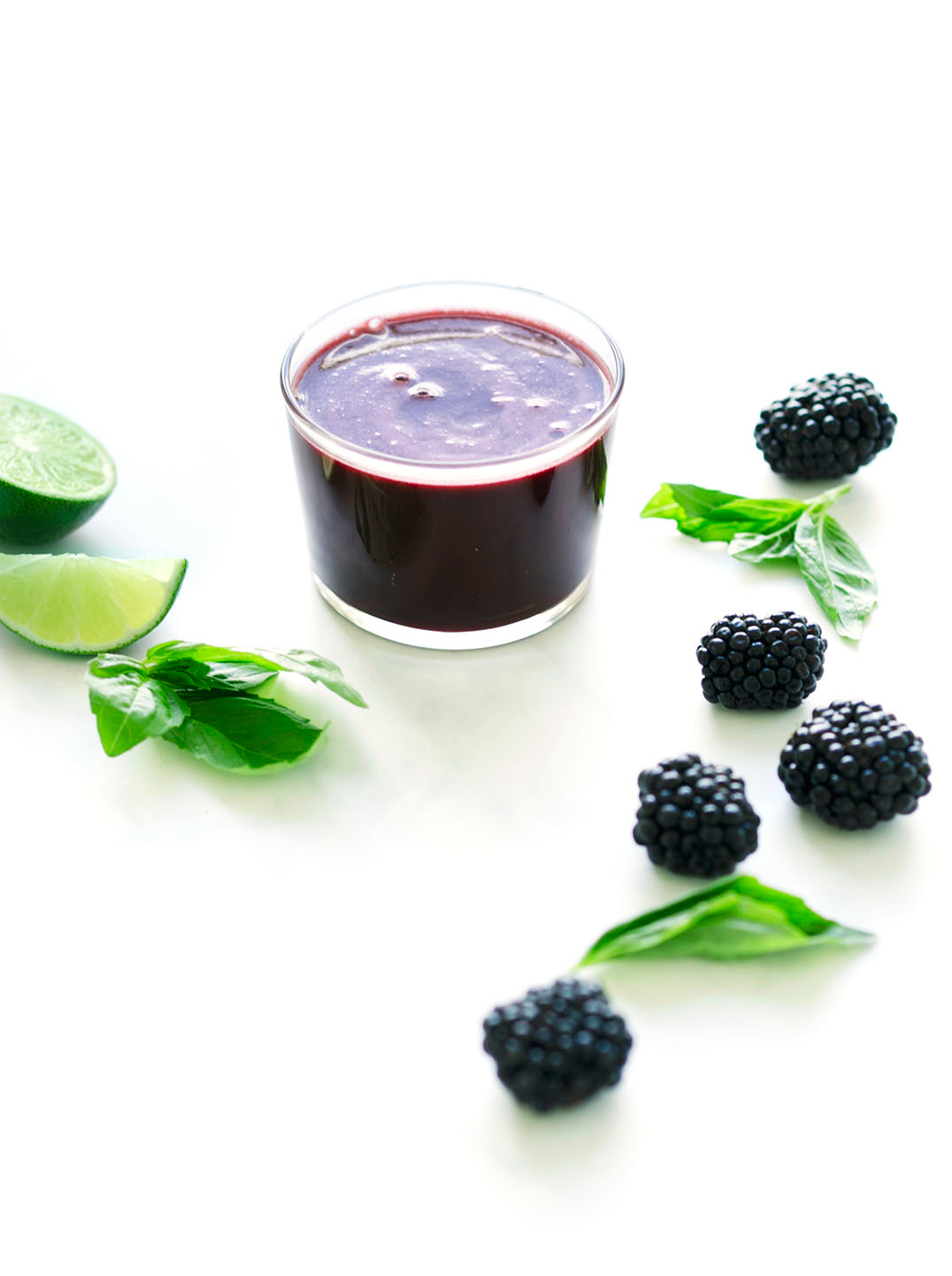 A glass of dark purple juice surrounded by ingredients blackberries lime and basil
