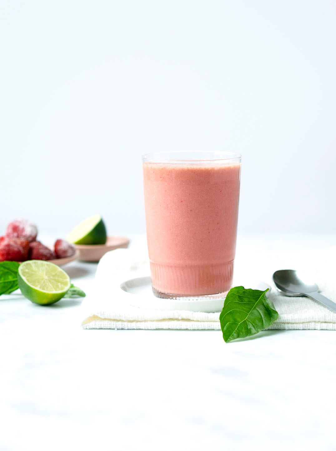 A glass filled with a beautiful pink smoothie surrounded by frozen strawberries, cut limes, and a basil leaf 