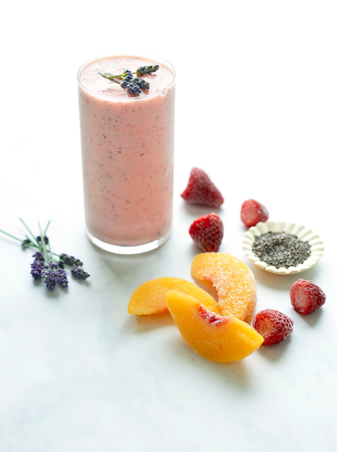 A tall glass filled with a pink smoothie surrounded by ingredients including peach strawberries and lavendar