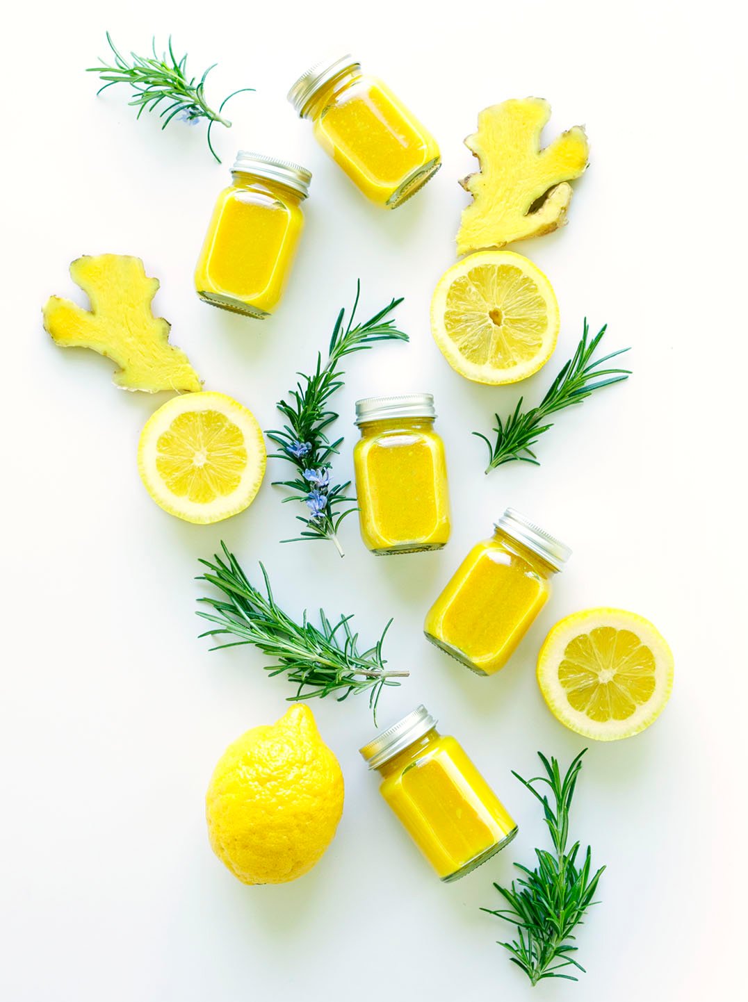 Overhead view of 5 small bottles filled with yellow juice surrounded by sprigs of rosemary lemon and ginger slices.