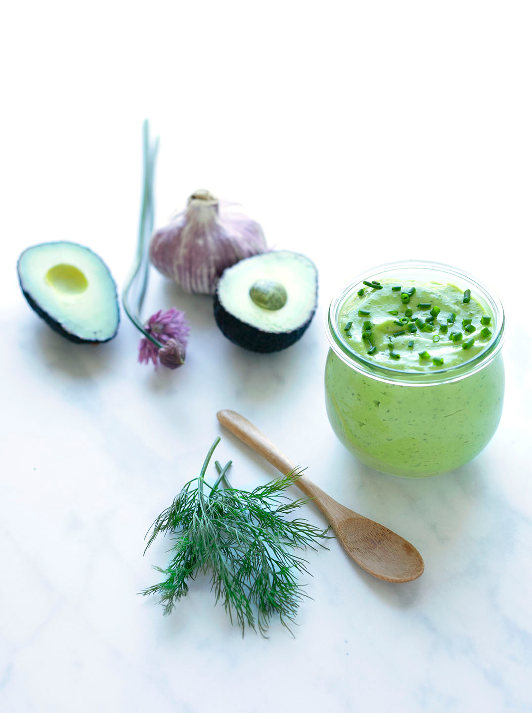A jar of creamy green dressing topped with chive 2 halves of an avocado garlic dill a chive flower and wooden spoon