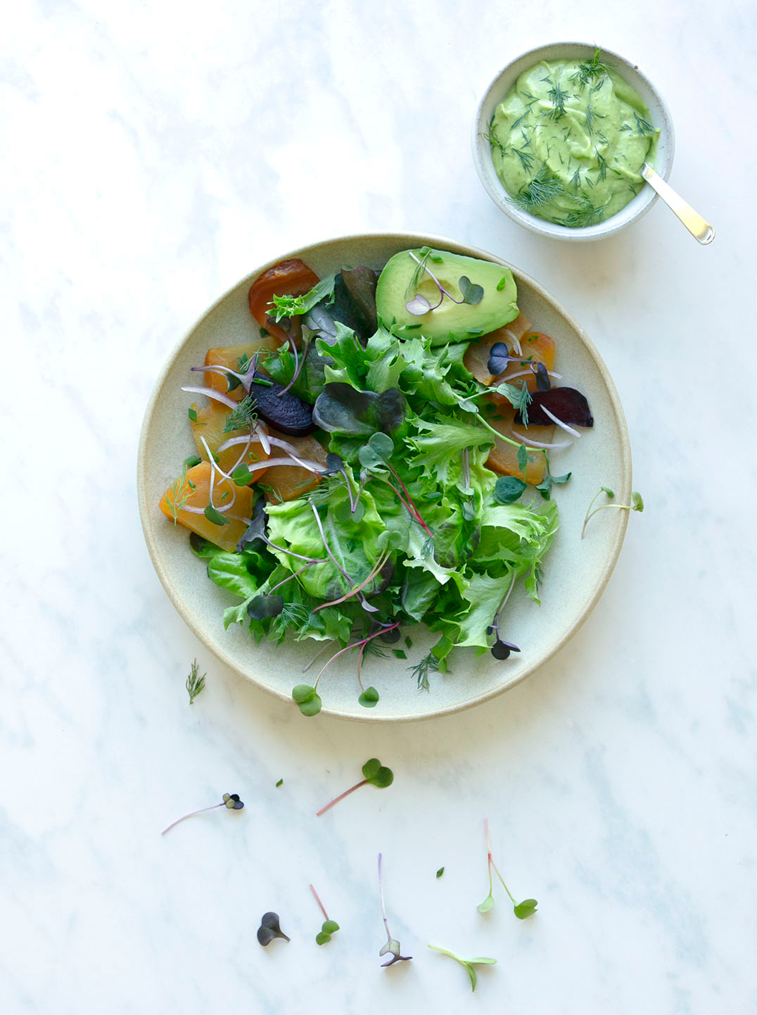 A bowl of microgreens with half an avocado roasted yellow beets and herbs on a marble surface