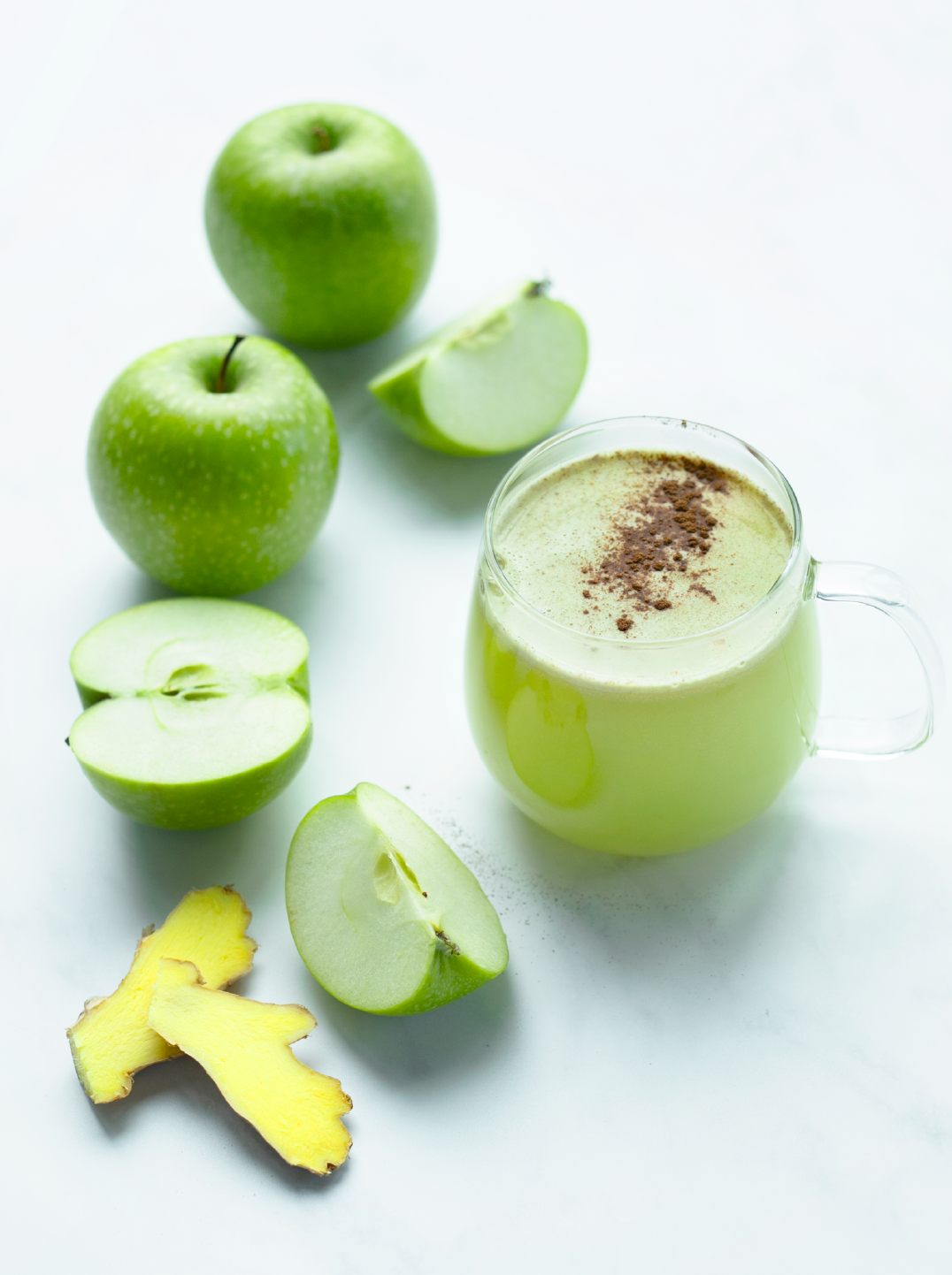 A glass of cozy ginger applle juice surrounded by green apples and a slice of ginger root 