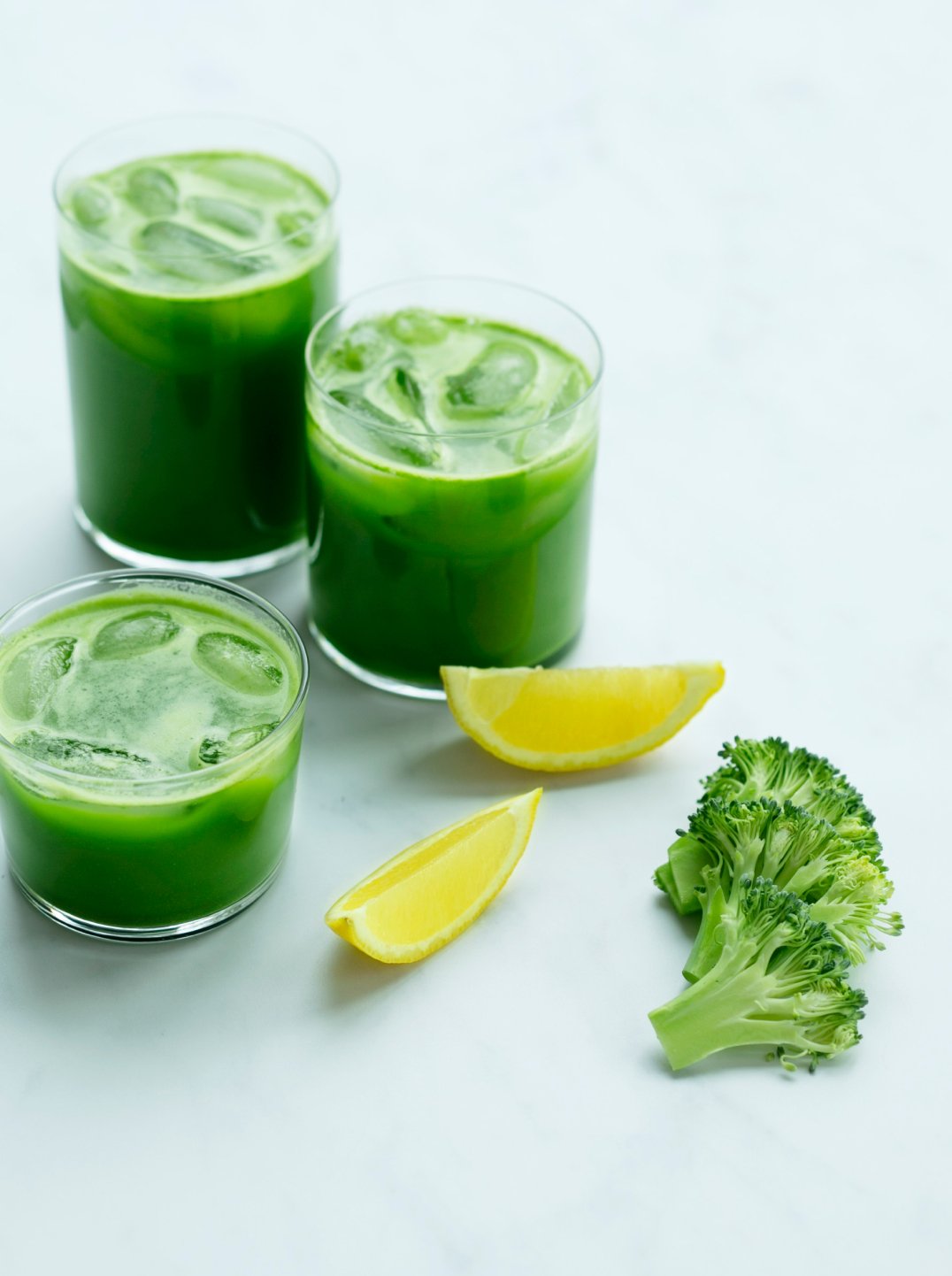 three glasses of green juice with lemon wedges and broccoli tips