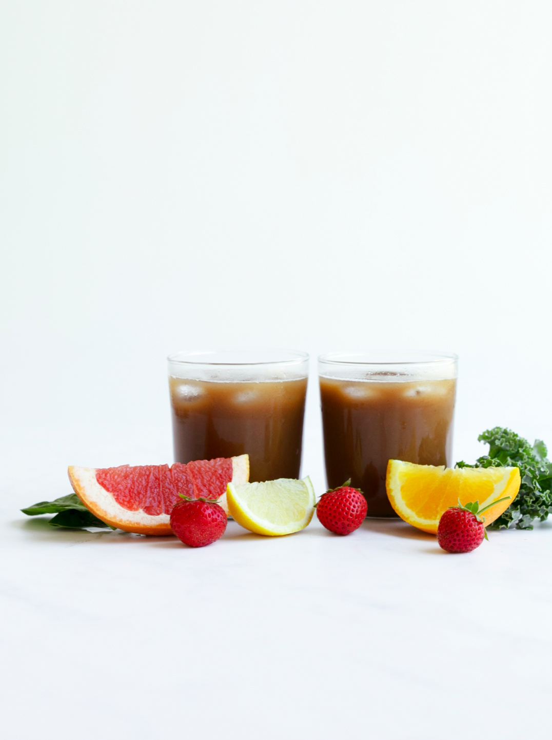 Two glasses of rusty red juice iced with grapefruit lemon and orange and strawberries  in front. 