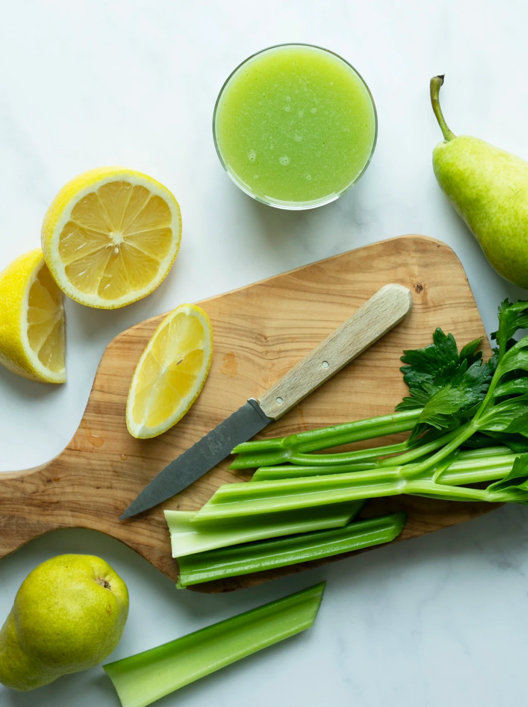 A glass of green juice with ingredients pear lemon and celery and cutting board