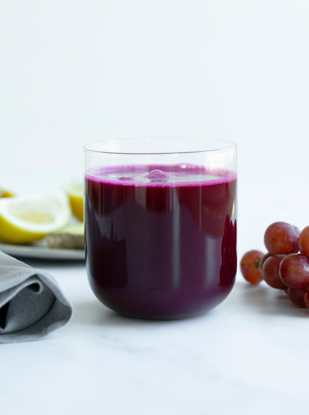 A round bottom glass filled with purple power juice on a marble counter with a piece of lemon and red grapes 