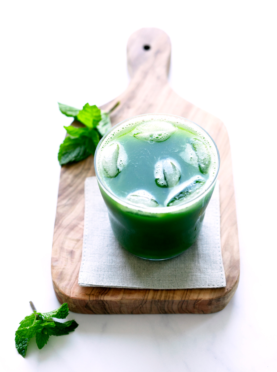 A glass filled with mint green juice with ice on a wooden cutting board with a sprig of mint