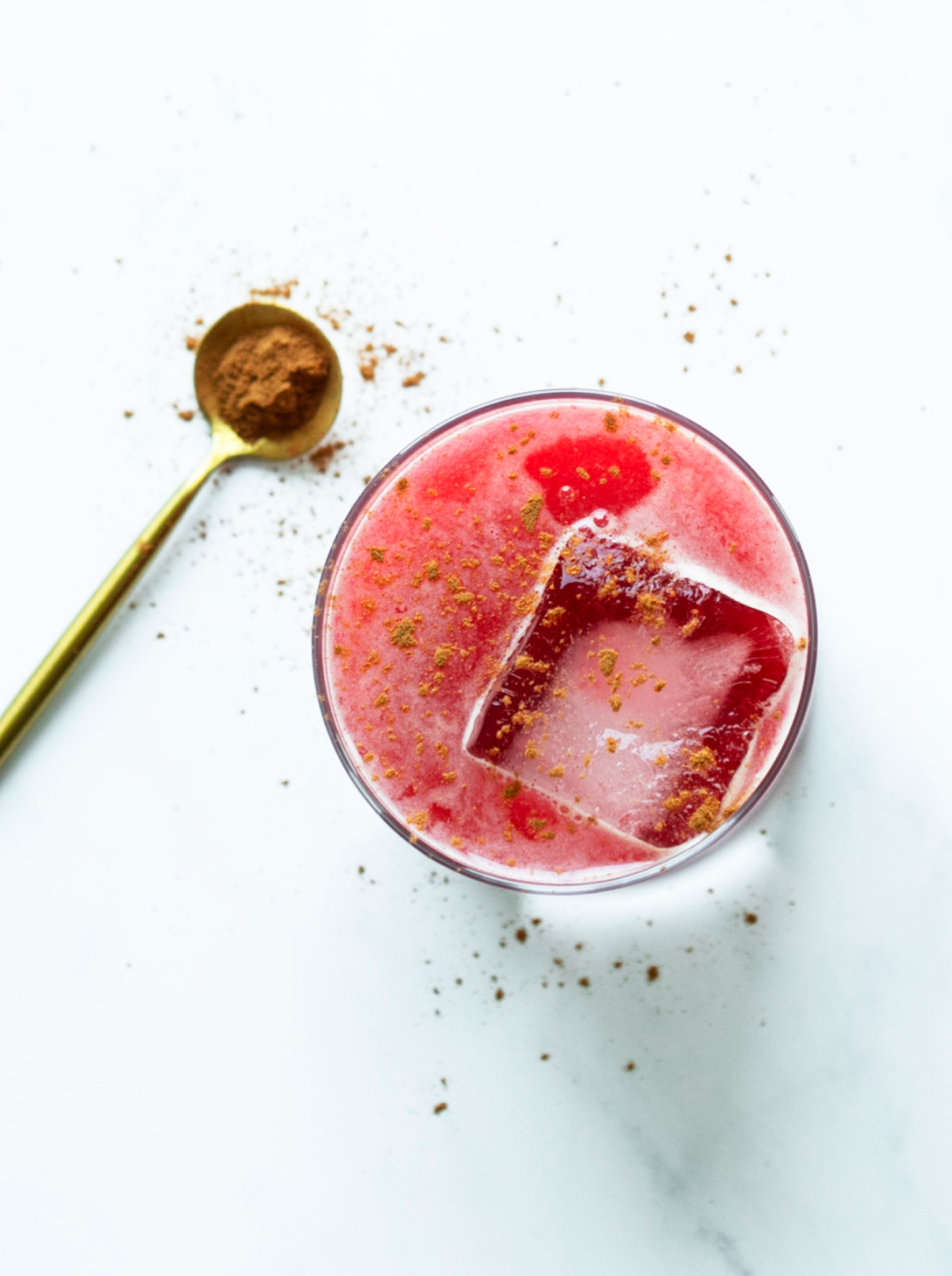 A glass of pink Cran-Apple Crisp Juice with ice and a spoonful of cinnamon