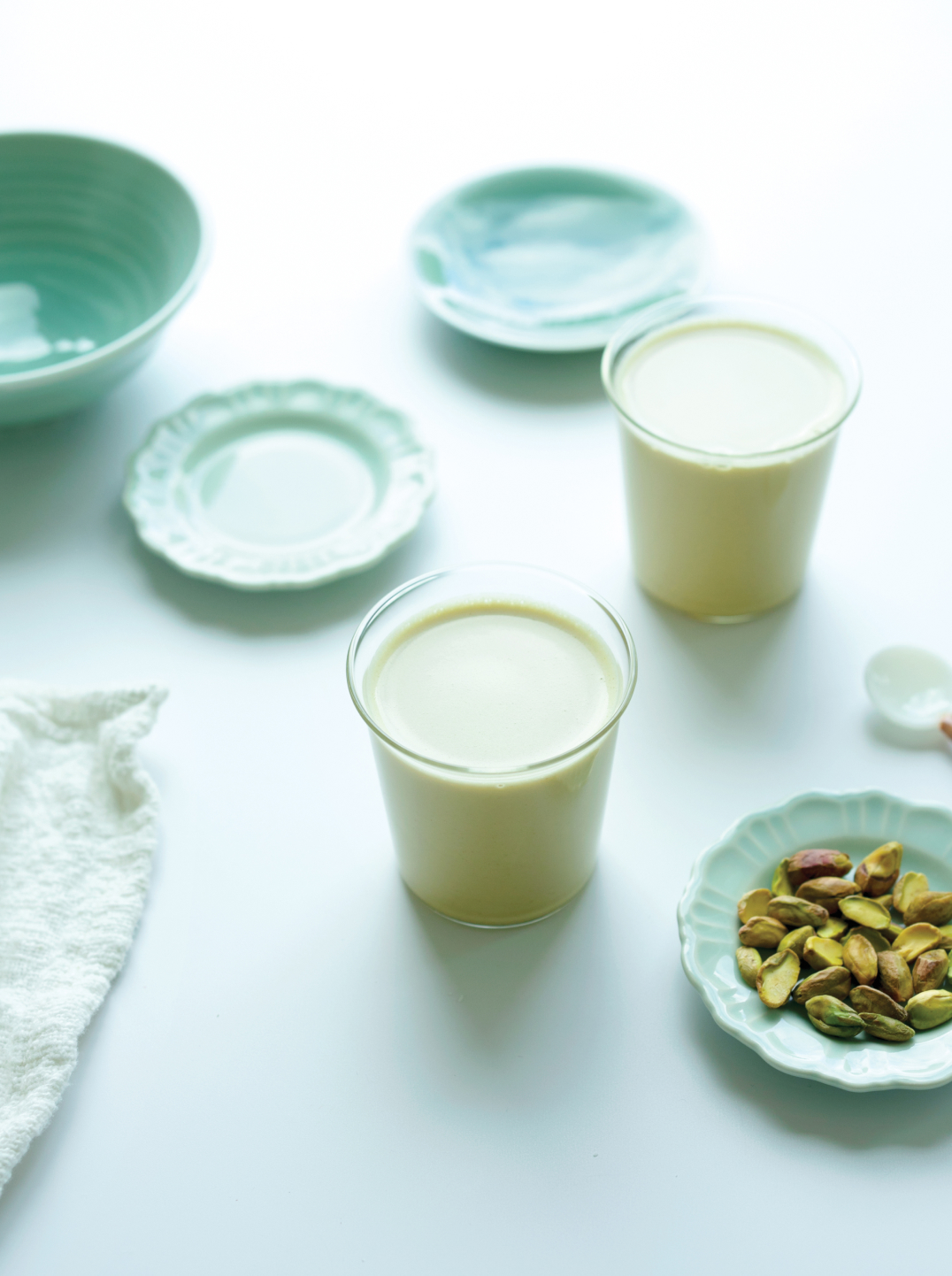 Two glasses of Pistachio Matcha Milk on a counter with 3 green plates one with pistachios and a bowl on a marble surface